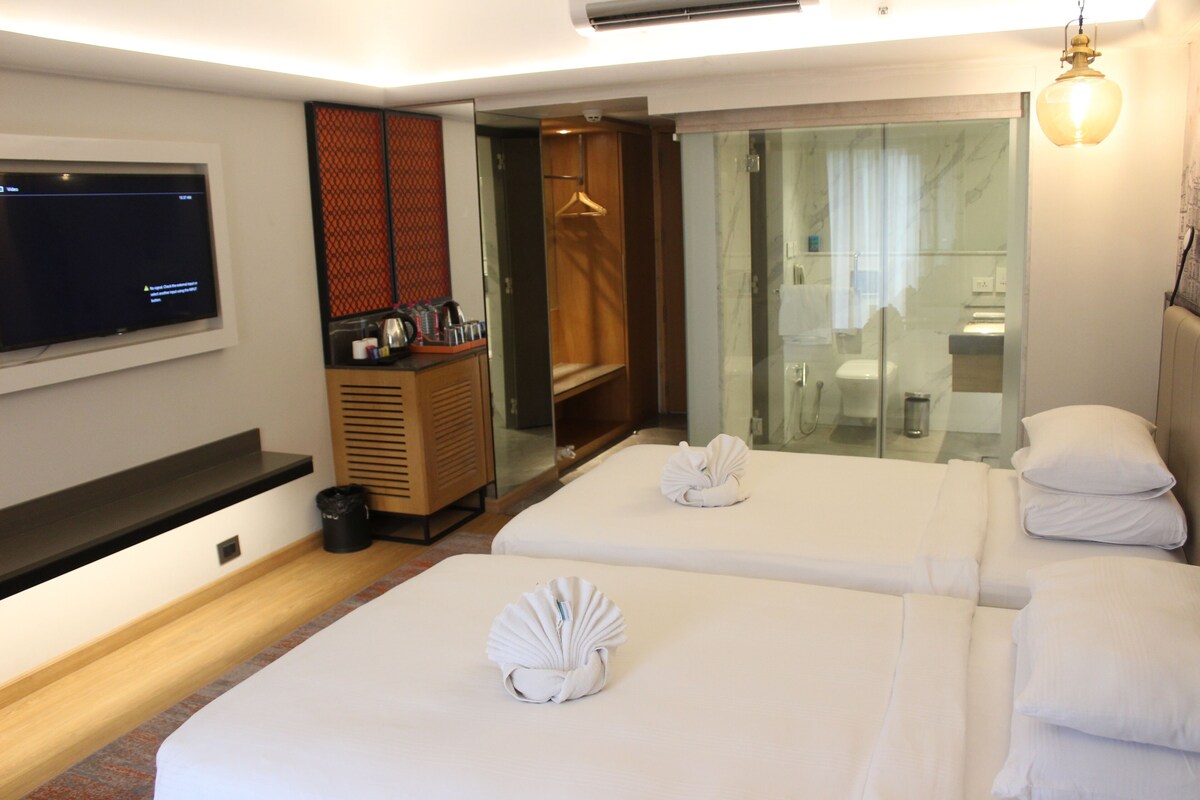 A hotel room with two beds, a TV, a minibar, a bathroom visible through a glass door, and a hanging light fixture.