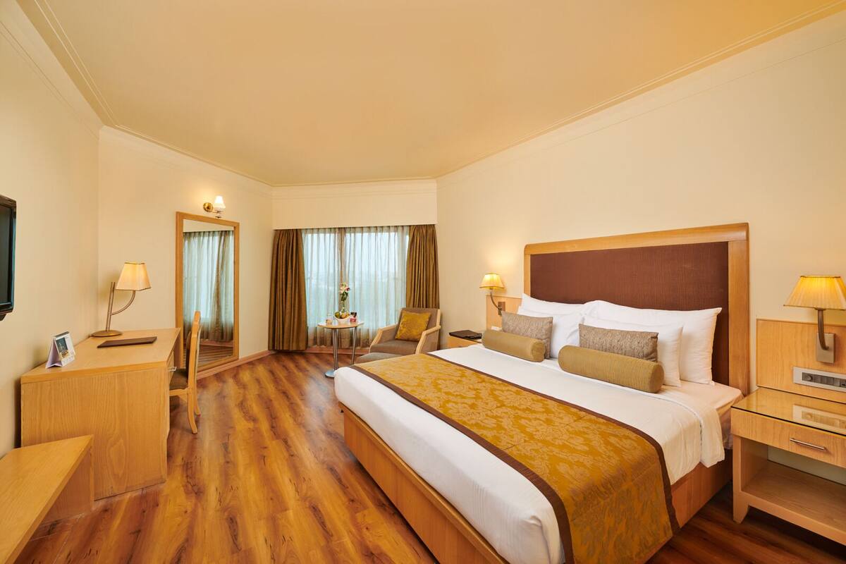 Ein Hotelzimmer mit einem großen Bett, einem Schreibtisch, einem Sessel, einem kleinen Tisch mit Lampe und einem Fernseher an der Wand.