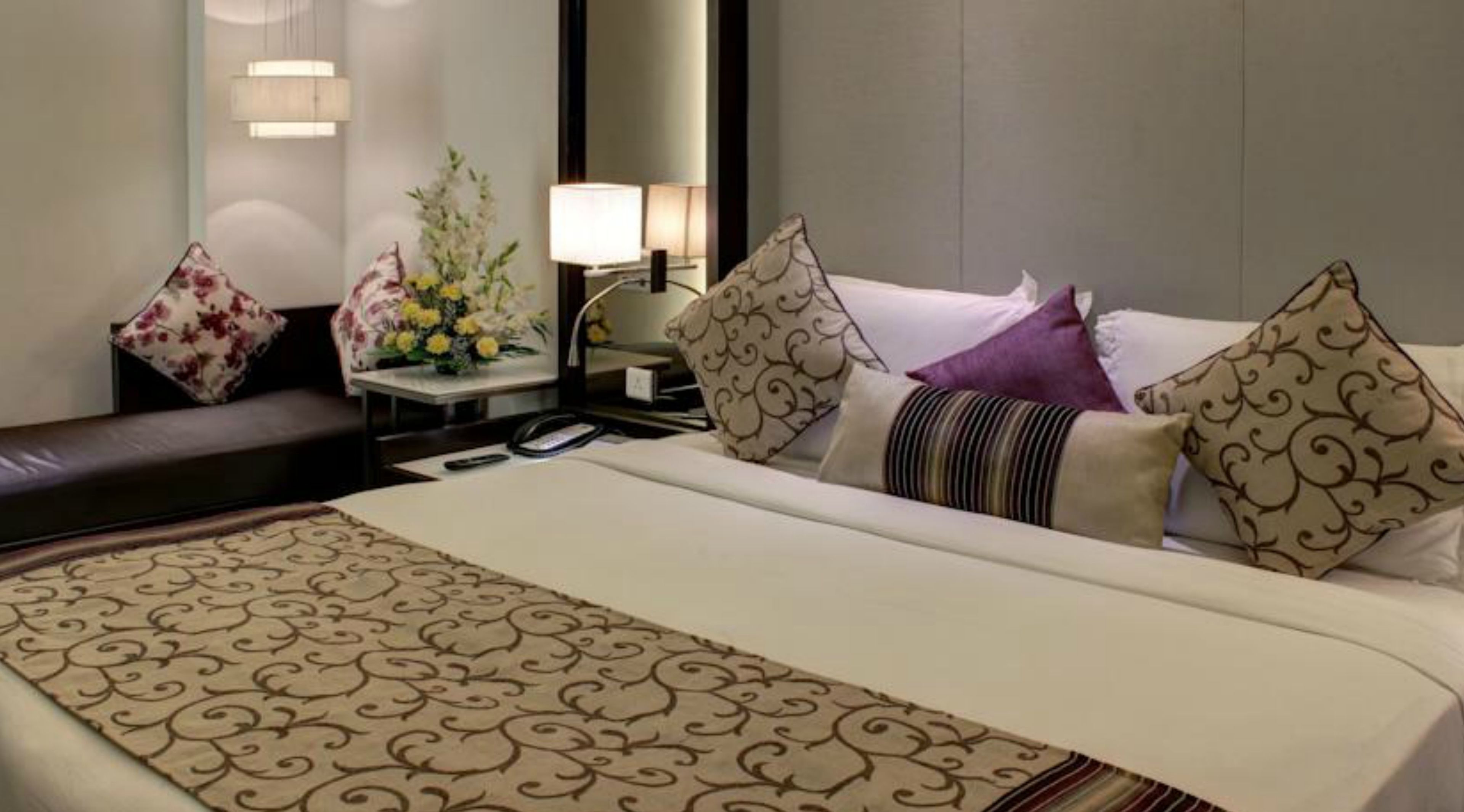 Une chambre d’hôtel avec un grand lit, une table de chevet avec une lampe, un arrangement floral et un banc en cuir marron.