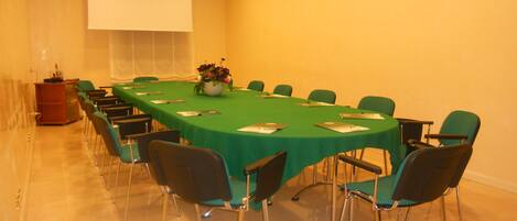 Un salón de conferencias con una larga mesa, sillas verdes y una pantalla para proyector.
