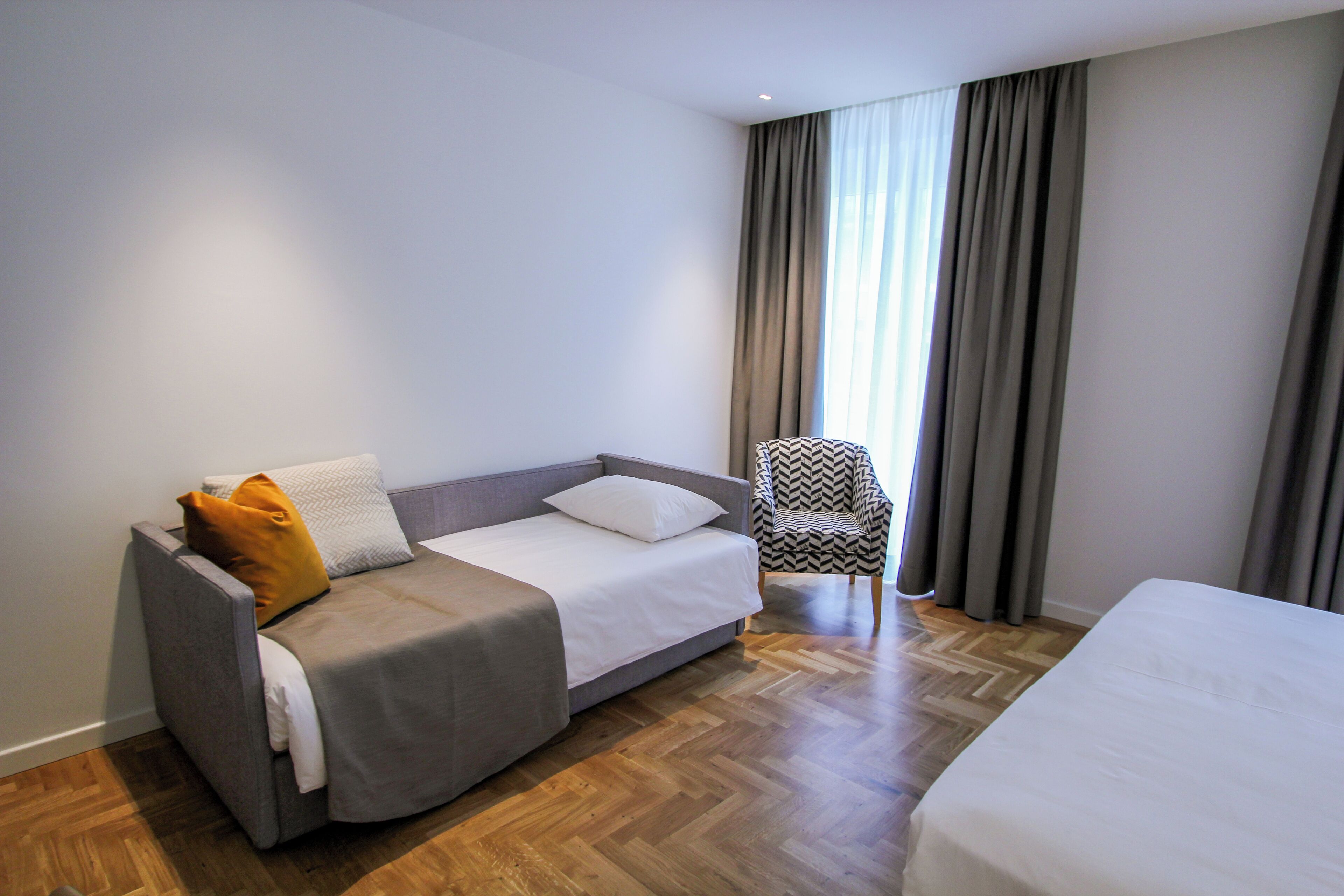 A modern hotel room with a grey sofa, a patterned armchair, and a large bed.