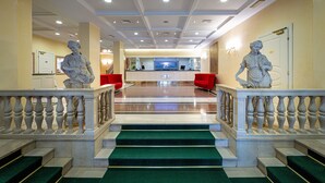 A grand staircase with green carpeted steps leading to a reception area with a TV and red chairs. - Ambassador Palace Hotel (Udine)