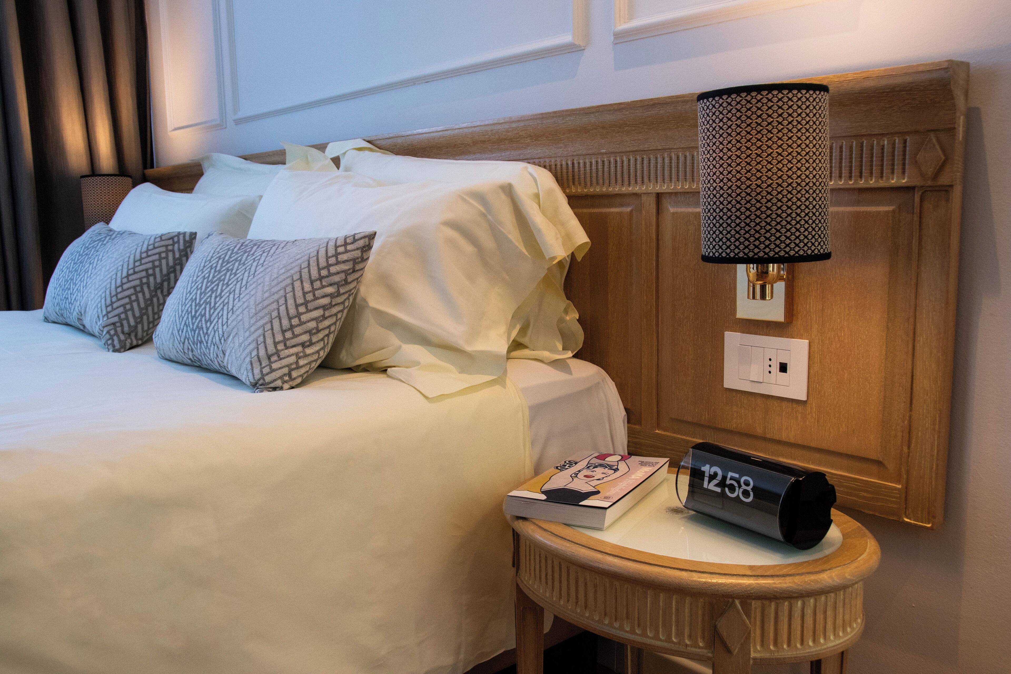 Una cama bien tendida con sábanas blancas y almohadas estampadas, una mesita de noche con un libro y un reloj, y una lámpara de pared.