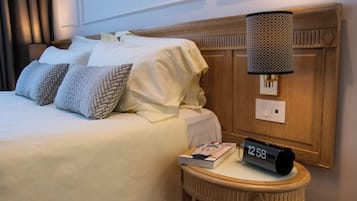 Un letto rifatto con lenzuola bianche e cuscini decorati, un comodino con un libro e una sveglia, e una lampada a parete.