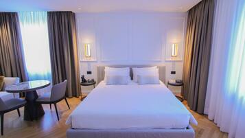 A modern hotel room with a large bed, a round dining table, two chairs, and a small side table with a clock.