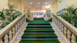 A grand staircase with green carpet and wooden balustrades leading to a reception area with a counter and decorative statues. - Ambassador Palace Hotel (Udine)
