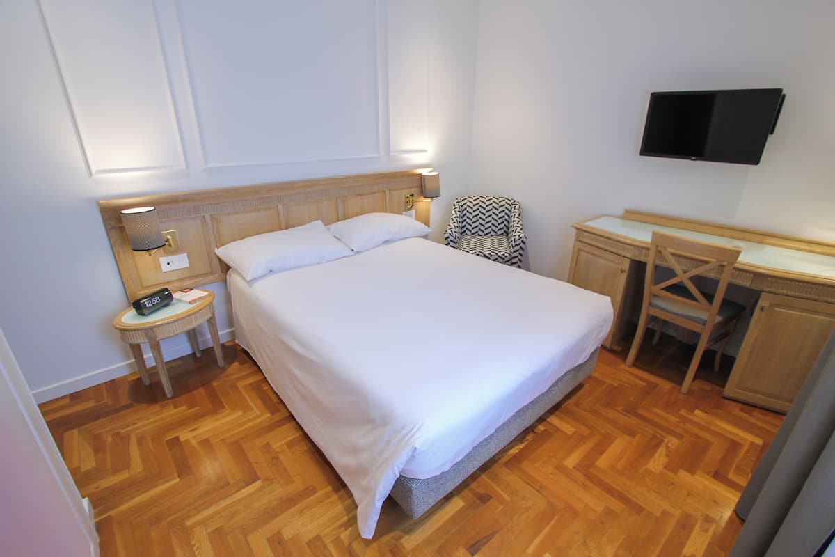 Un dormitorio ordenado con una cama grande, mesita de noche, silla, escritorio y un televisor de pantalla plana montado en la pared.