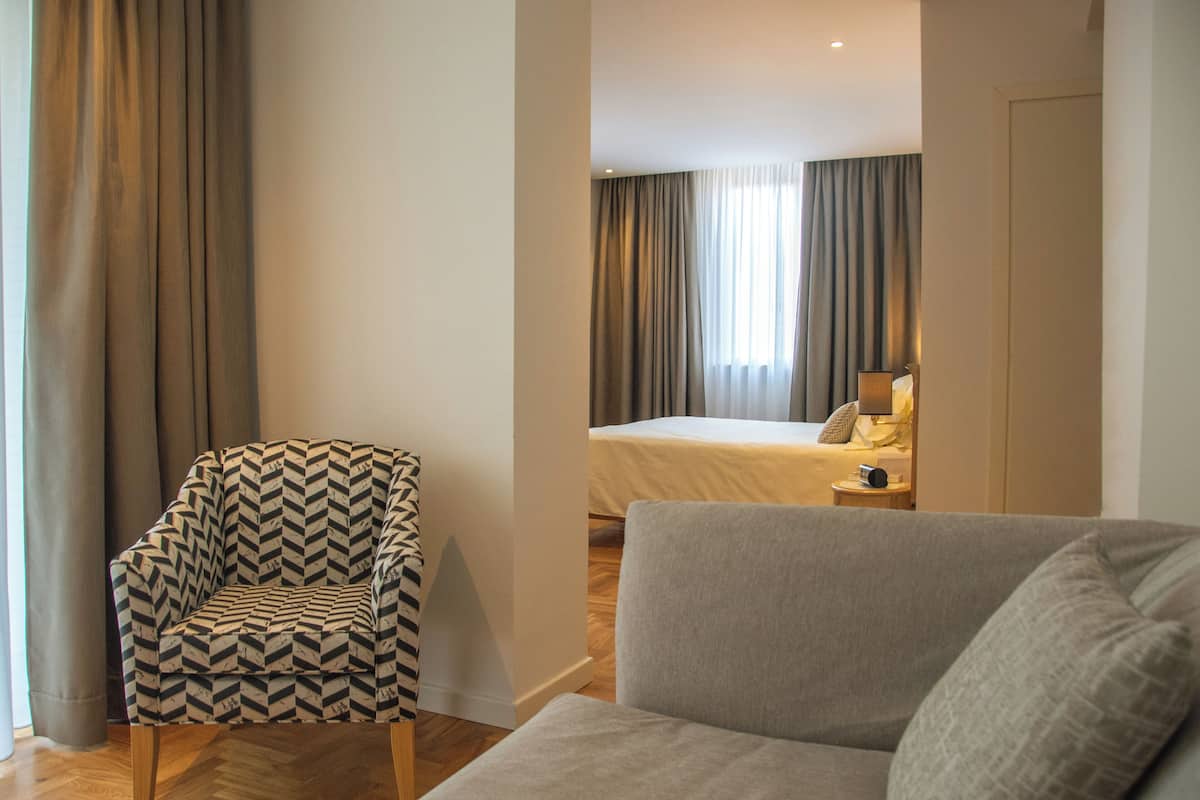 Una habitación de hotel con un sillón estampado, un sofá gris, una cama con edredón blanco y una mesita de noche con lámpara.