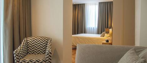 A hotel room with a patterned armchair, a grey sofa, a bed with a white duvet, and a bedside table with a lamp.