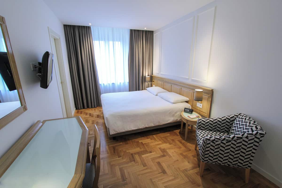 Habitación de hotel con una cama grande, una mesita de noche, una silla, un televisor montado en la pared y una ventana con cortinas.