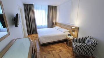 Une chambre d’hôtel avec un grand lit, une table de chevet, une chaise, un téléviseur fixé au mur et une fenêtre avec des rideaux.