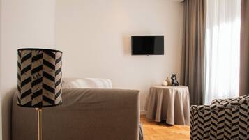 A hotel room with a bed, a TV mounted on the wall, a small table with a vase, and a patterned armchair.