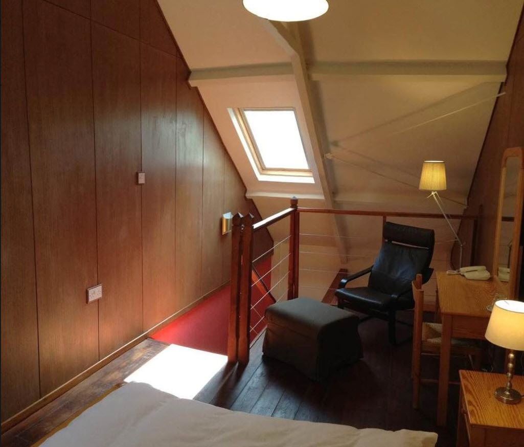 Una habitación con techo inclinado, paneles de madera, tragaluz, un escritorio con lámpara, una silla y una cama.