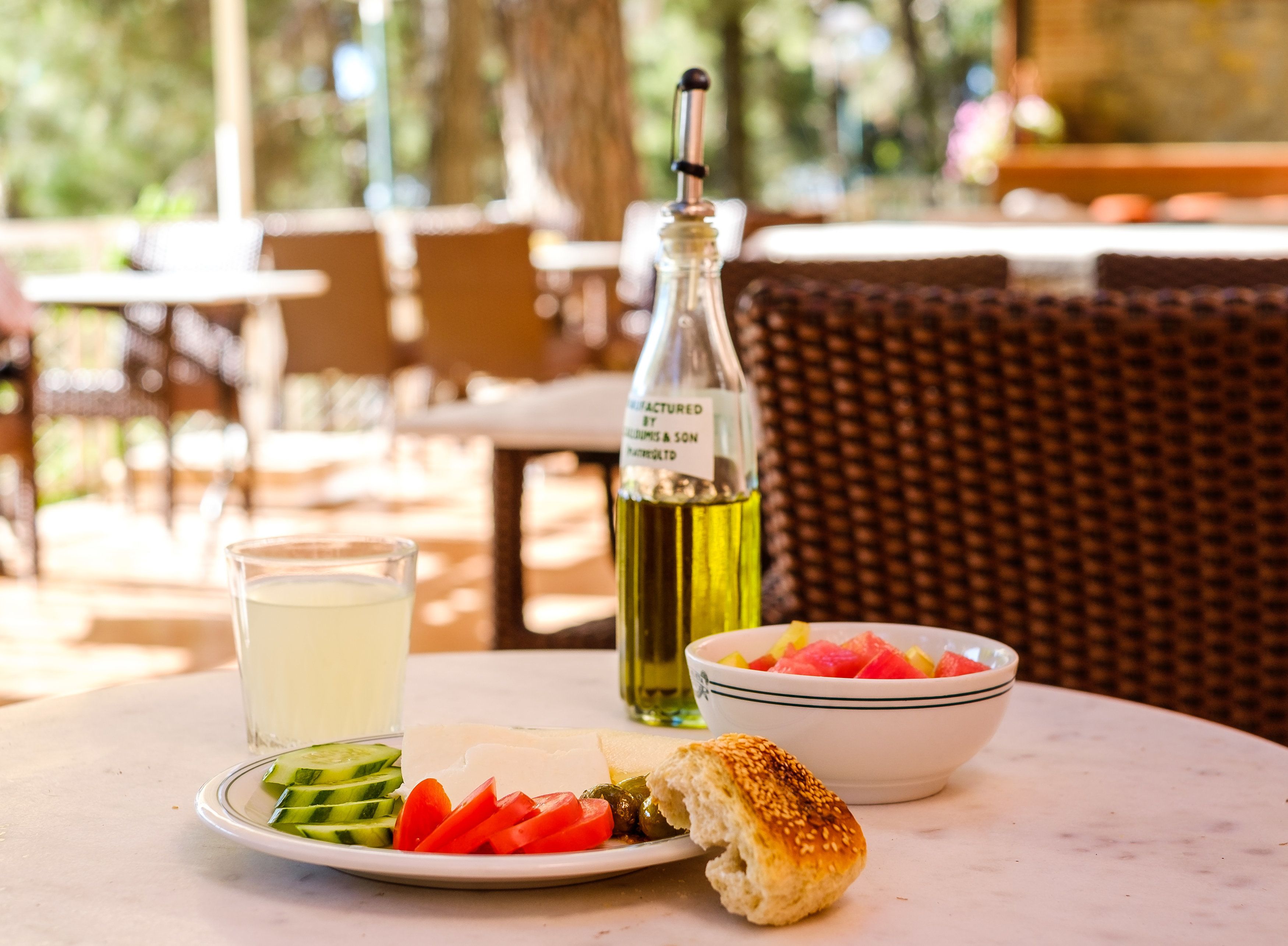 A table with a plate of sliced vegetables, a bowl of fruit, a bottle of olive oil, and a glass of water.