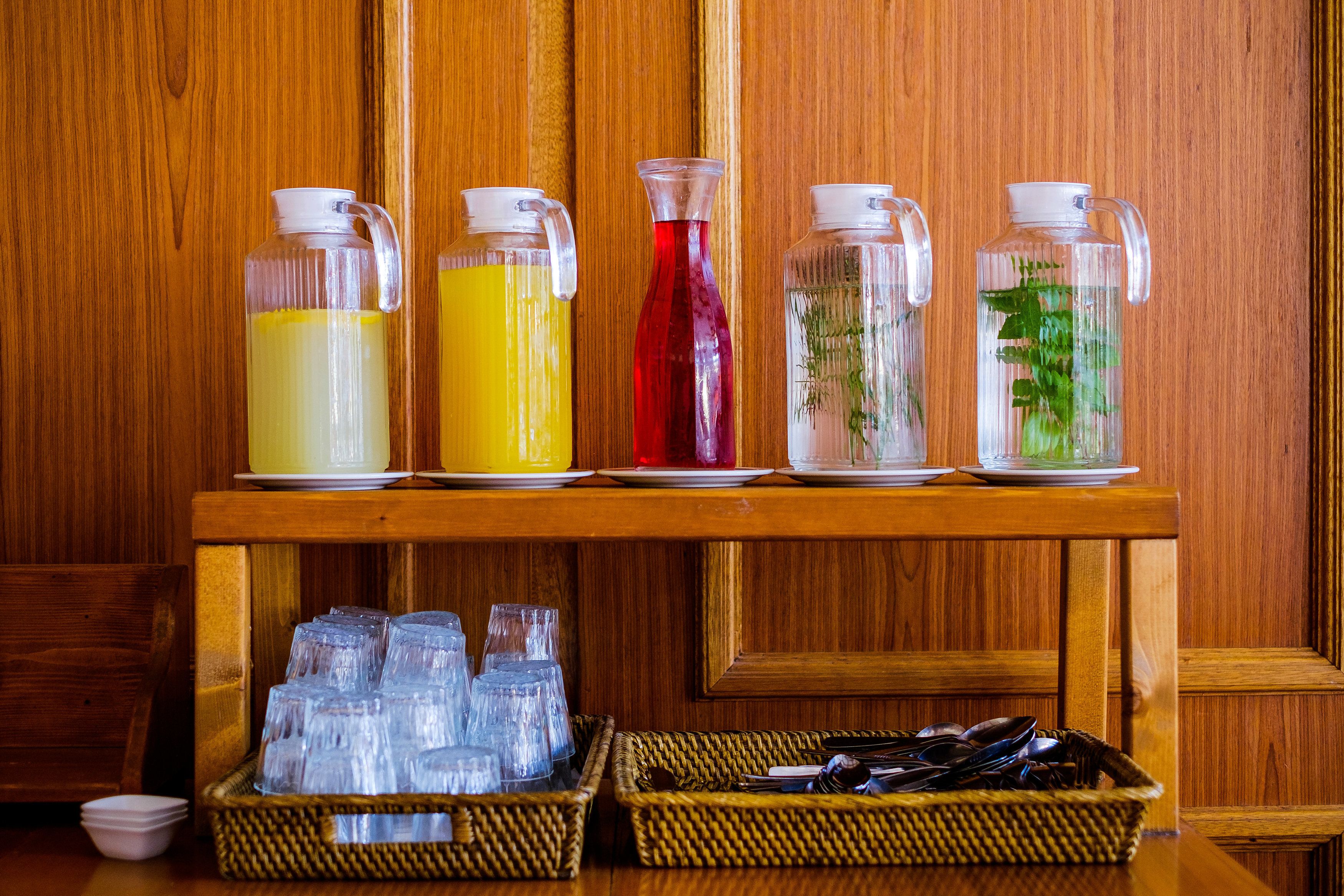 A wooden table with four glass pitchers of different beverages and two wicker baskets filled with clear plastic cups.