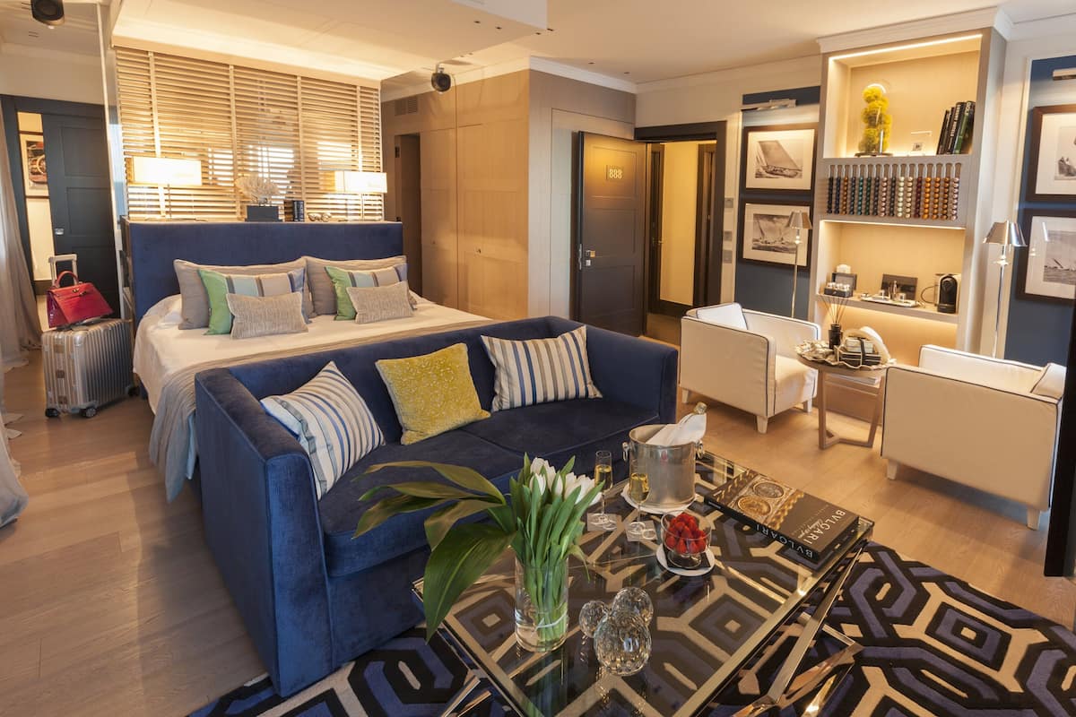 A modern hotel room with a large bed, a blue sofa, a coffee table, and a bookshelf.