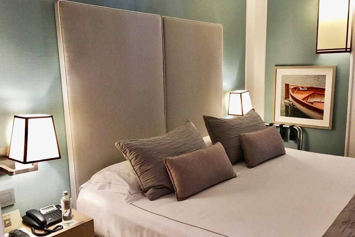 A hotel room with a large bed, two bedside lamps, a framed picture on the wall, and a nightstand with a phone and a bottle.