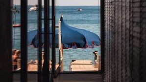 Vista desde una habitación con un ventanal grande que da a una playa con sombrillas y barcos.