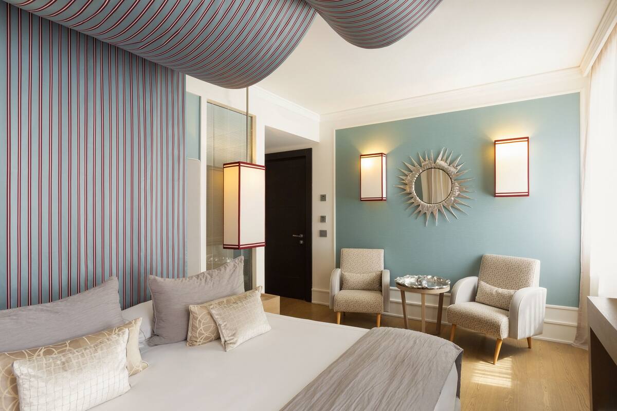 A modern hotel room with a large bed, two armchairs, a small table, and a mirror on the wall.