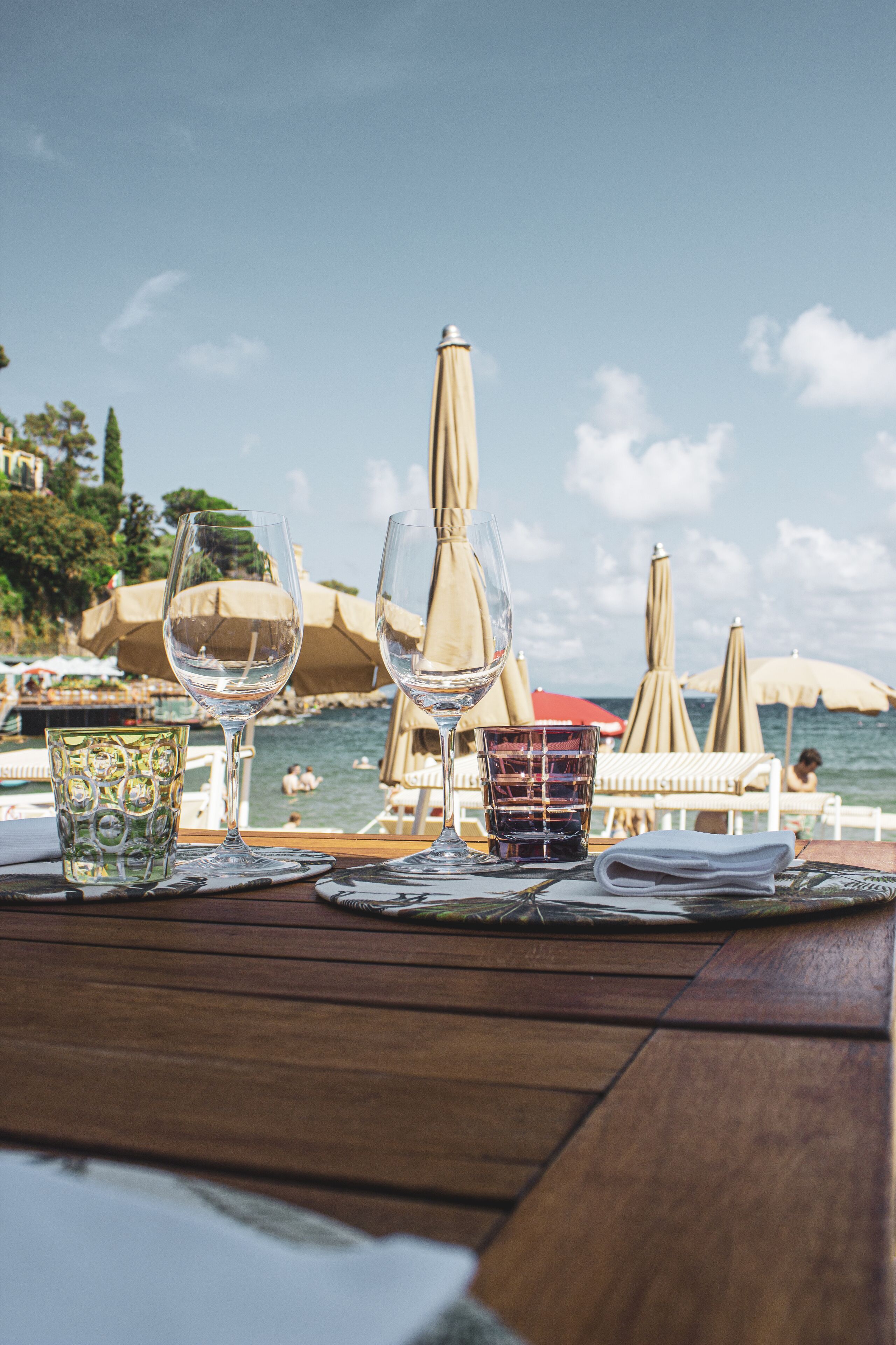 Ein Tisch mit Weingläsern und einer Speisekarte, mit Blick auf einen Strand mit Sonnenschirmen und Personen im Hintergrund.