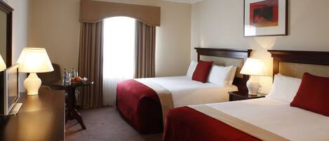 A hotel room with two beds, a desk with a lamp, a chair, and a window with curtains.