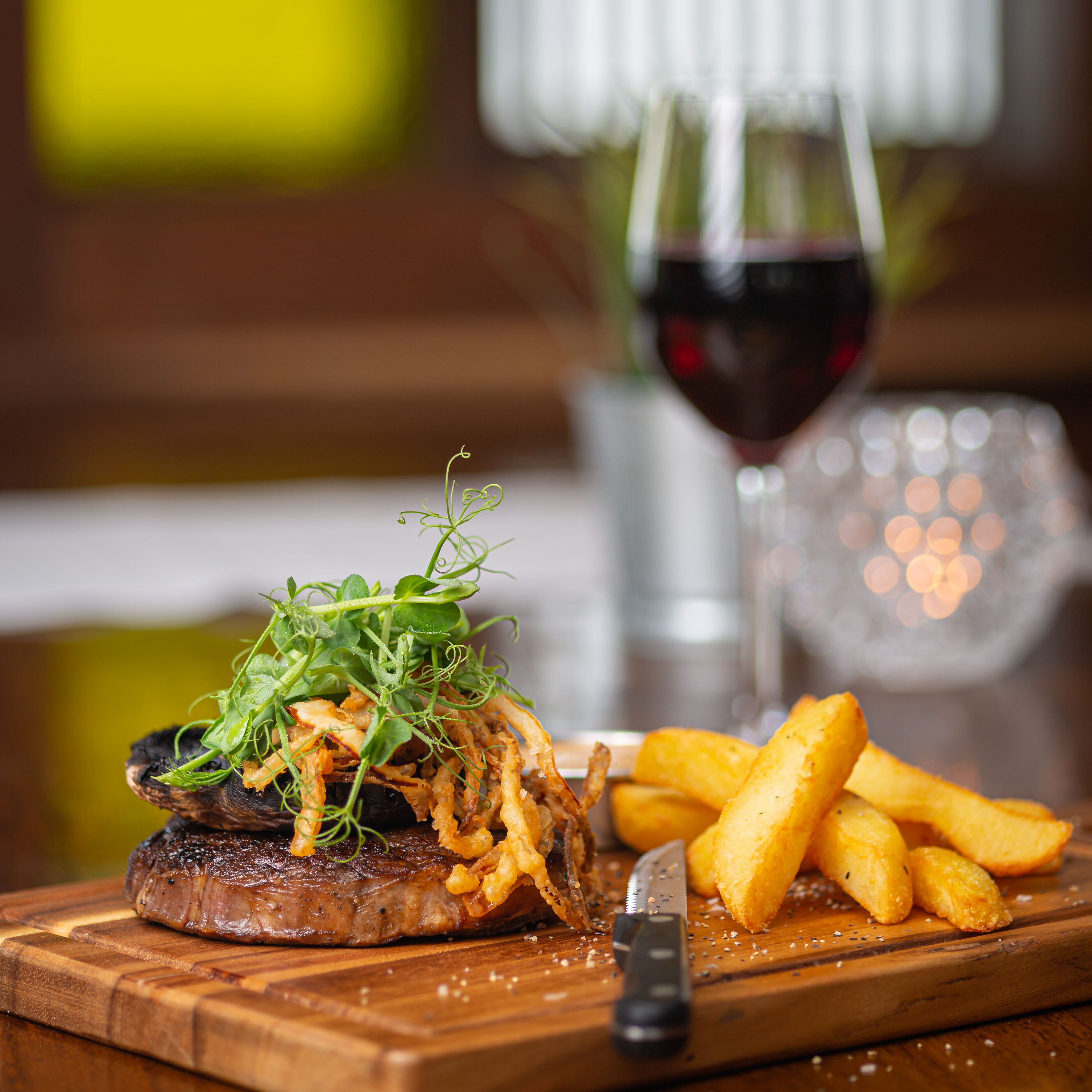 A steak with greens and fries on a wooden board, a knife, and a glass of red wine in the background.