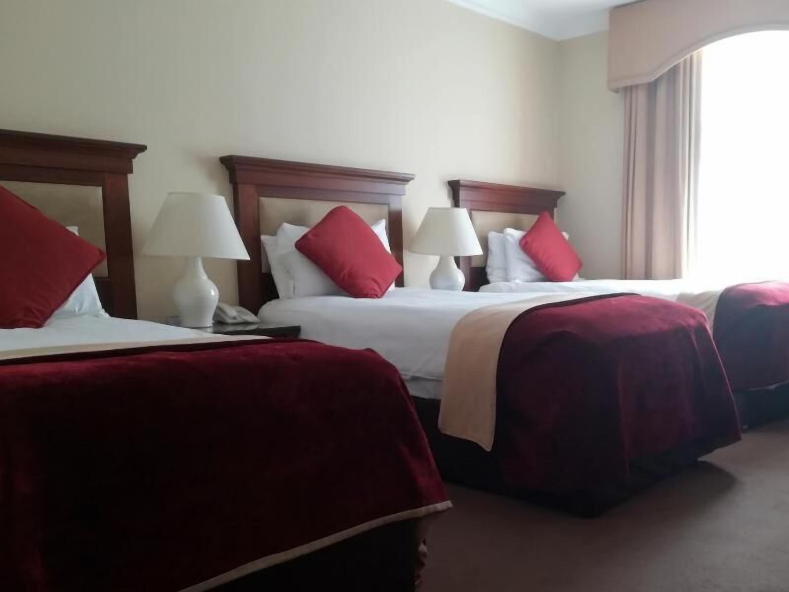 Habitación de hotel con tres camas, cada una con ropa de cama roja y almohadas blancas, y un ventanal con cortinas color beige.