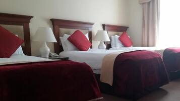 Habitación de hotel con tres camas, cada una con ropa de cama roja y almohadas blancas, y un ventanal con cortinas color beige.