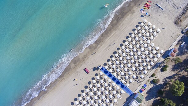 Vista aérea de una playa con filas de sombrillas y sillas, algunas personas y un pequeño bote cerca del agua.