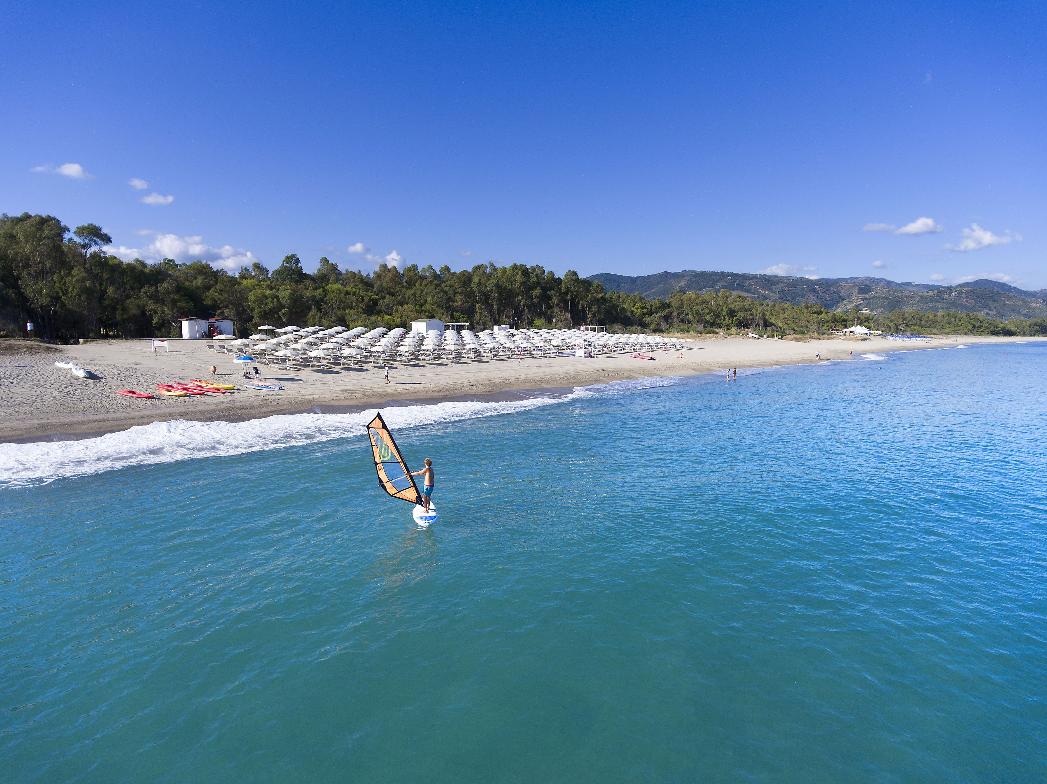 Ein Strand mit blauem Himmel, eine Person beim Windsurfen und Reihen von Strandstühlen.