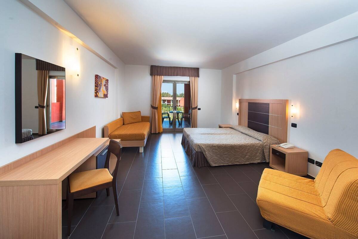 Habitación de hotel con cama, escritorio, silla, mesita de noche, espejo y vistas al exterior.