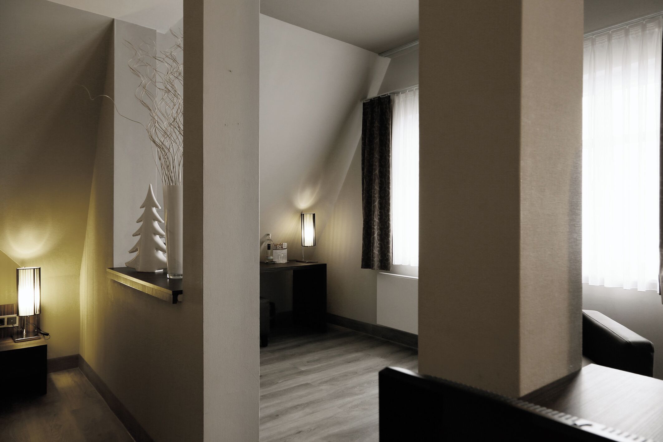 A modern hotel room with a wooden floor, a desk with a lamp, a small plant, and a window with curtains.