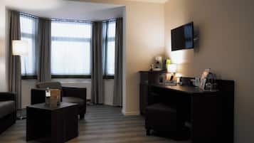 A hotel room with a dark wood floor, a desk with a lamp, a television on the wall, and a view of the outside through large windows with curtains.