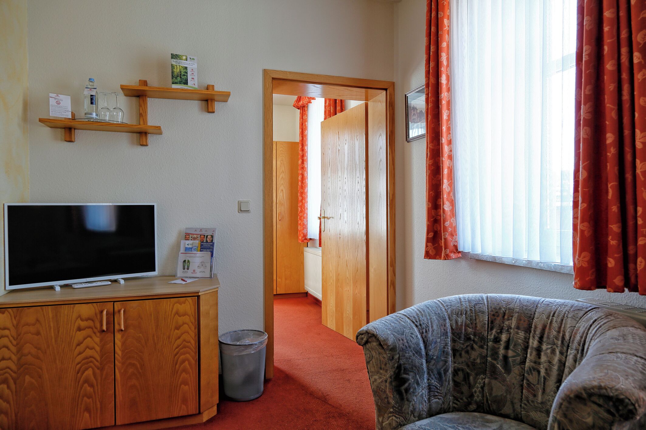 A room with a television on a wooden cabinet, a patterned sofa, a hallway leading to a door, and a window with curtains.