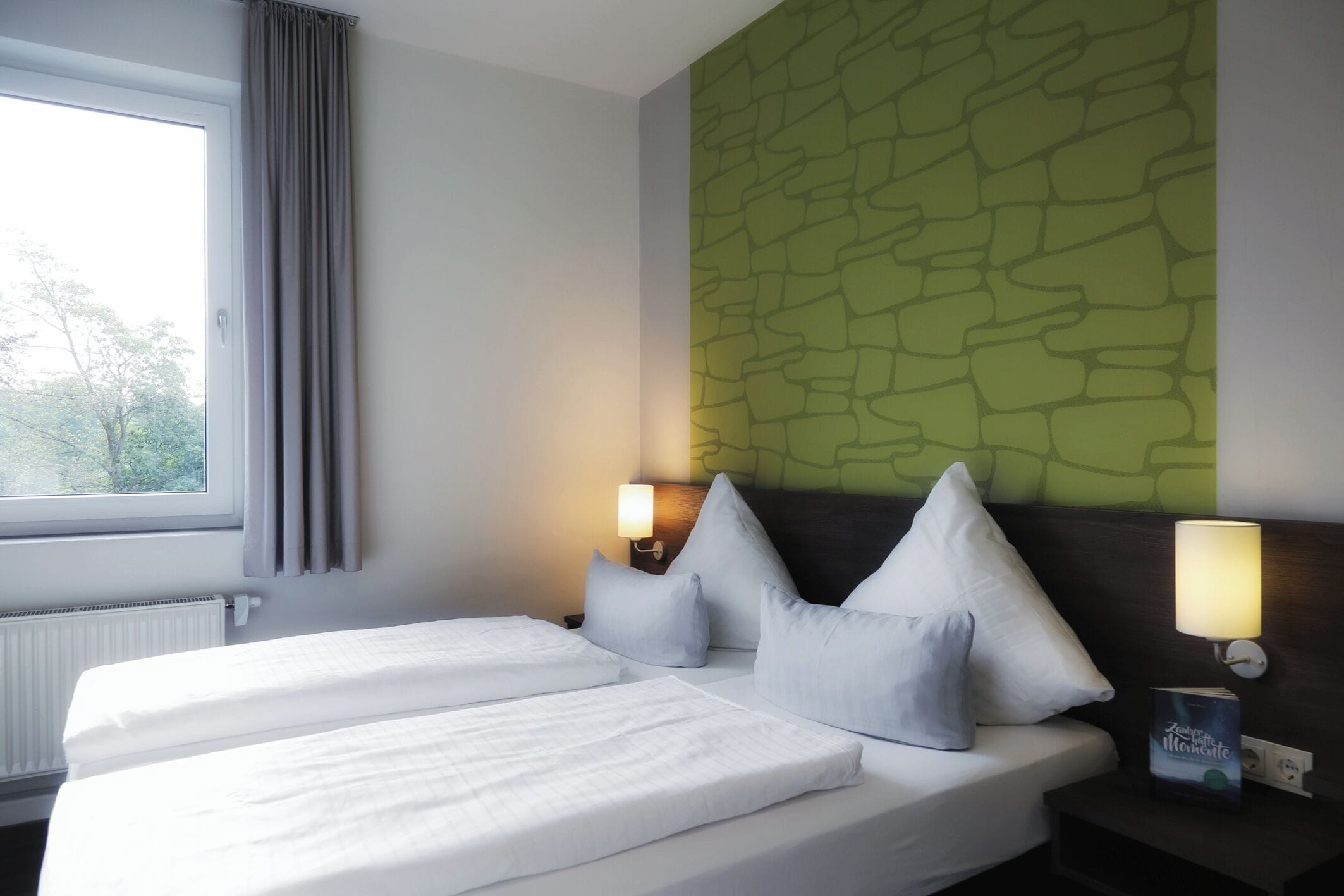 A hotel room with two beds, a window with curtains, a green textured wall, and two bedside lamps.