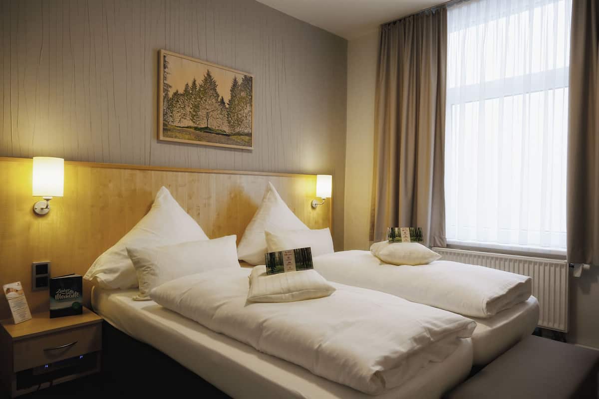 Une chambre d’hôtel avec deux lits, une table de chevet, une lampe, un tableau au mur et une fenêtre avec des rideaux.