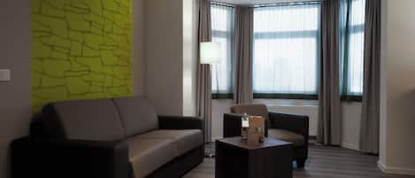 A modern living room with a sofa, a coffee table, and a lamp. There is a large window with curtains and a view of the outside.