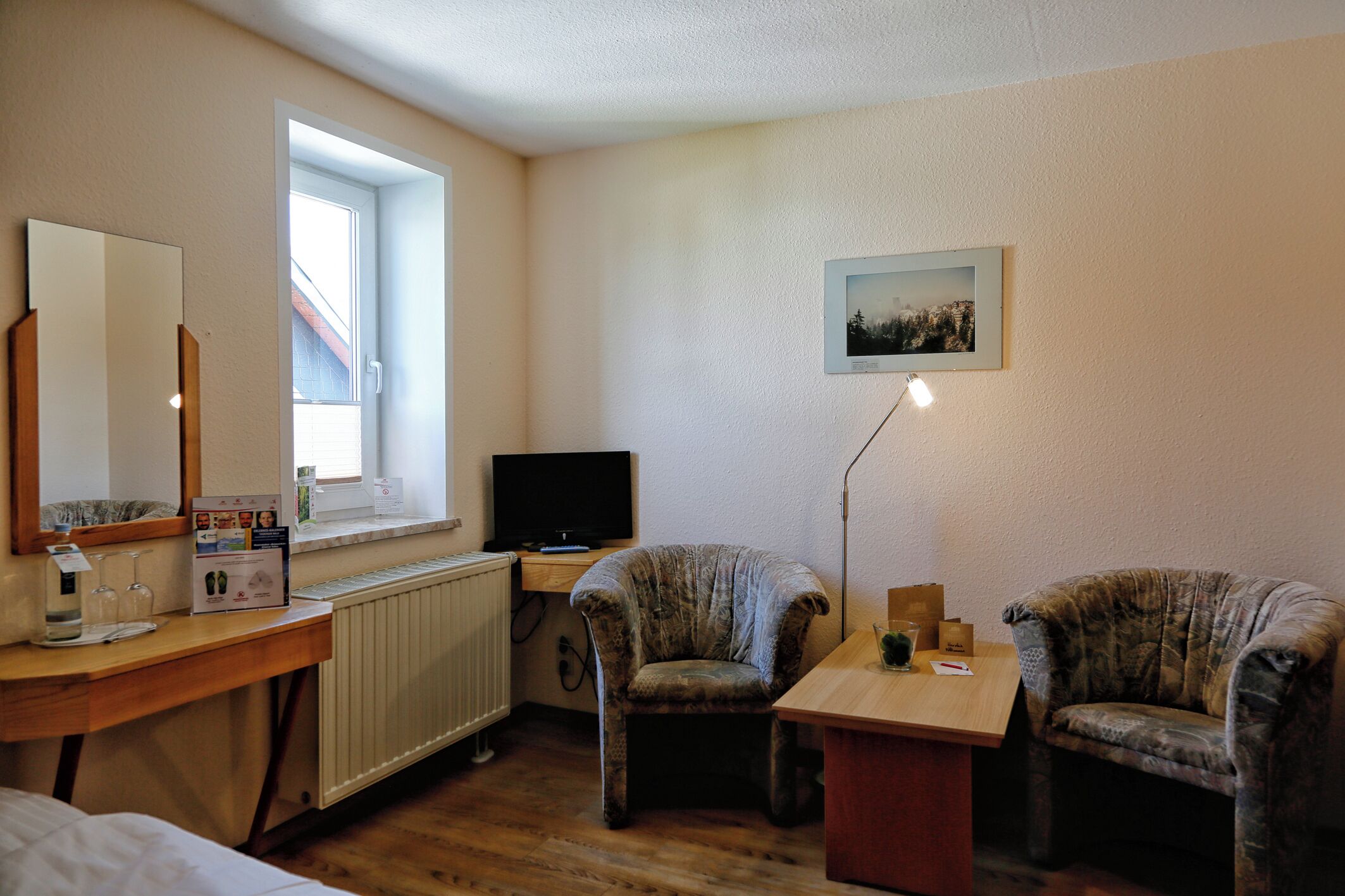 Una camera d'albergo con una scrivania in legno, una televisione, due poltrone, un tavolino, uno specchio, una finestra e un quadro appeso al muro.