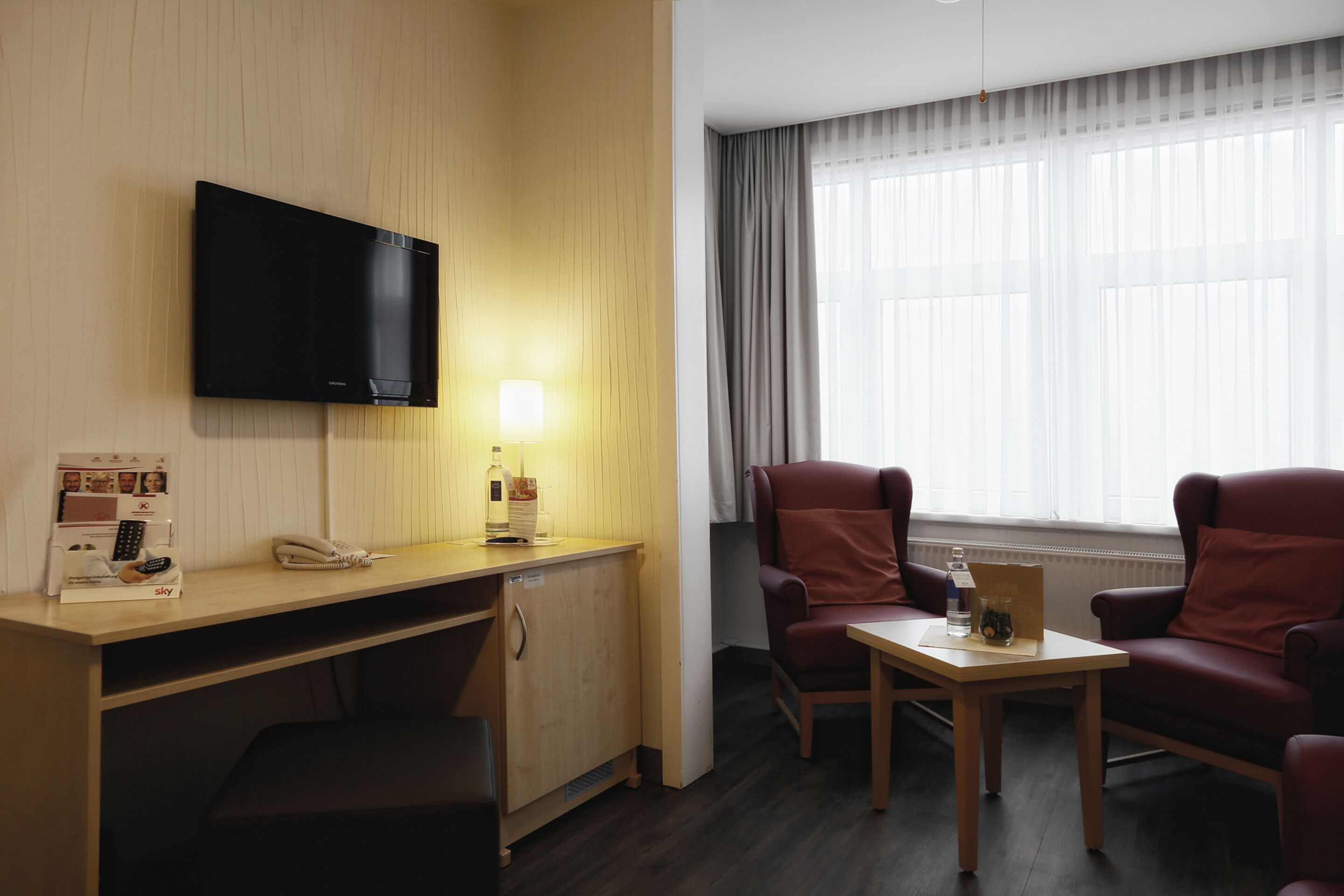 Een hotelkamer met een houten bureau, een flatscreen televisie aan de muur, een tafeltje met een lamp, twee rode fauteuils en een raam met gordijnen.