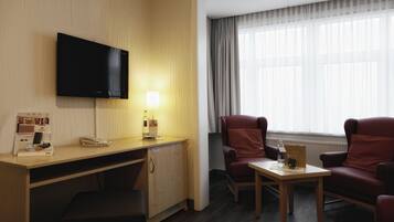 A hotel room with a wooden desk, a flat-screen TV mounted on the wall, a small table with a lamp, two red armchairs, and a window with curtains.