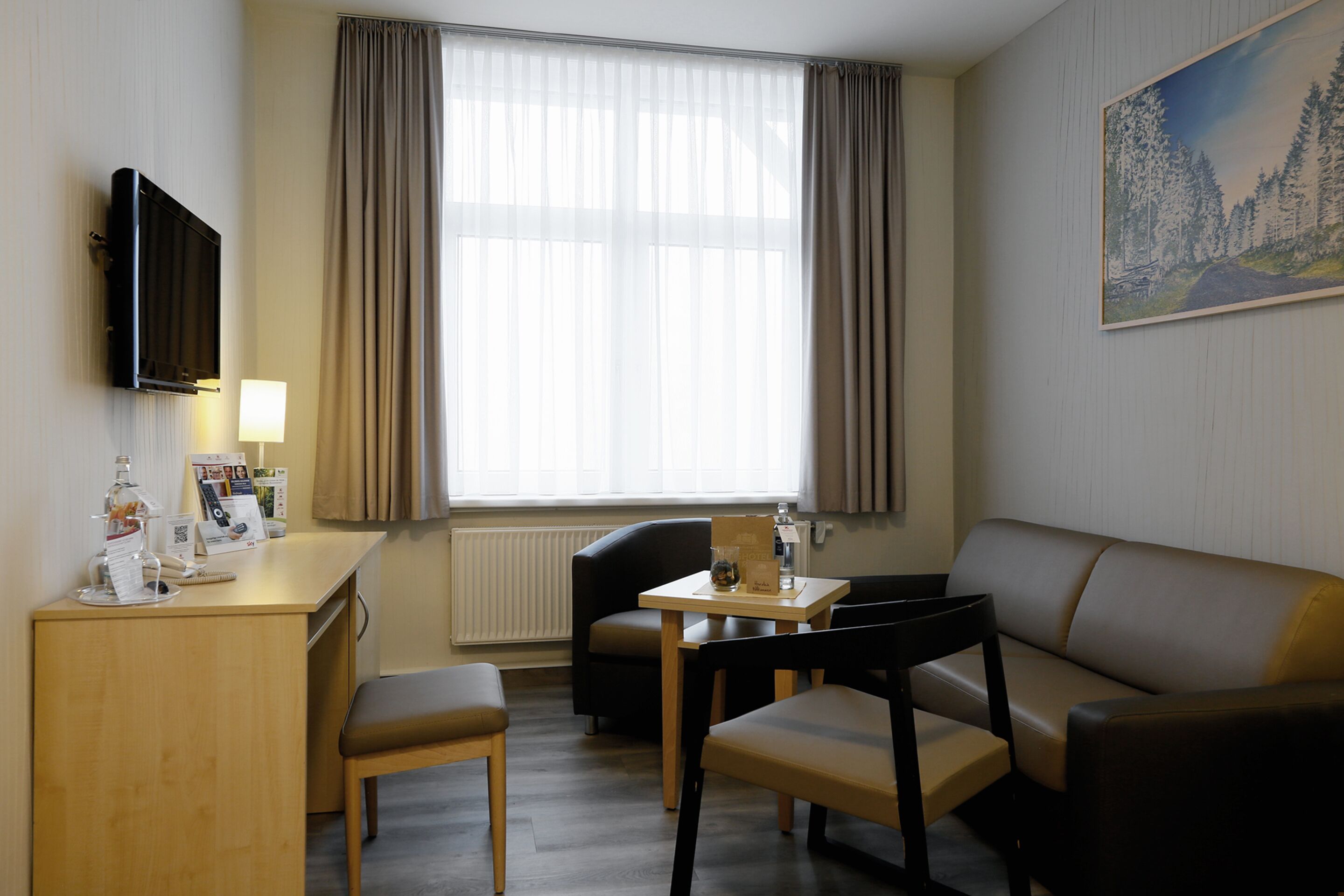 A hotel room with a wooden desk, a television, a sofa, and a large window with curtains.