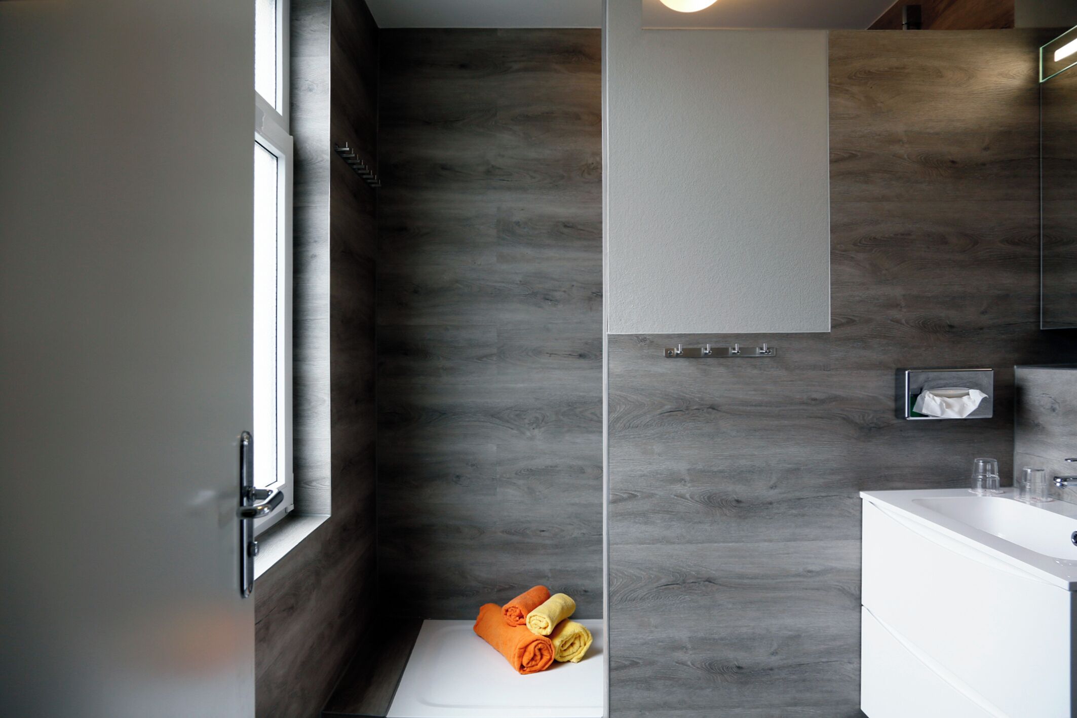 Een moderne badkamer met een groot raam, een witte wastafel en een houten wand.