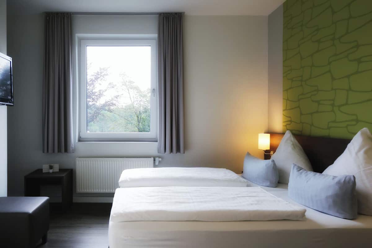 Une chambre d’hôtel avec deux lits, une fenêtre avec des rideaux, un radiateur, une table de chevet, une lampe et un mur vert à texture.