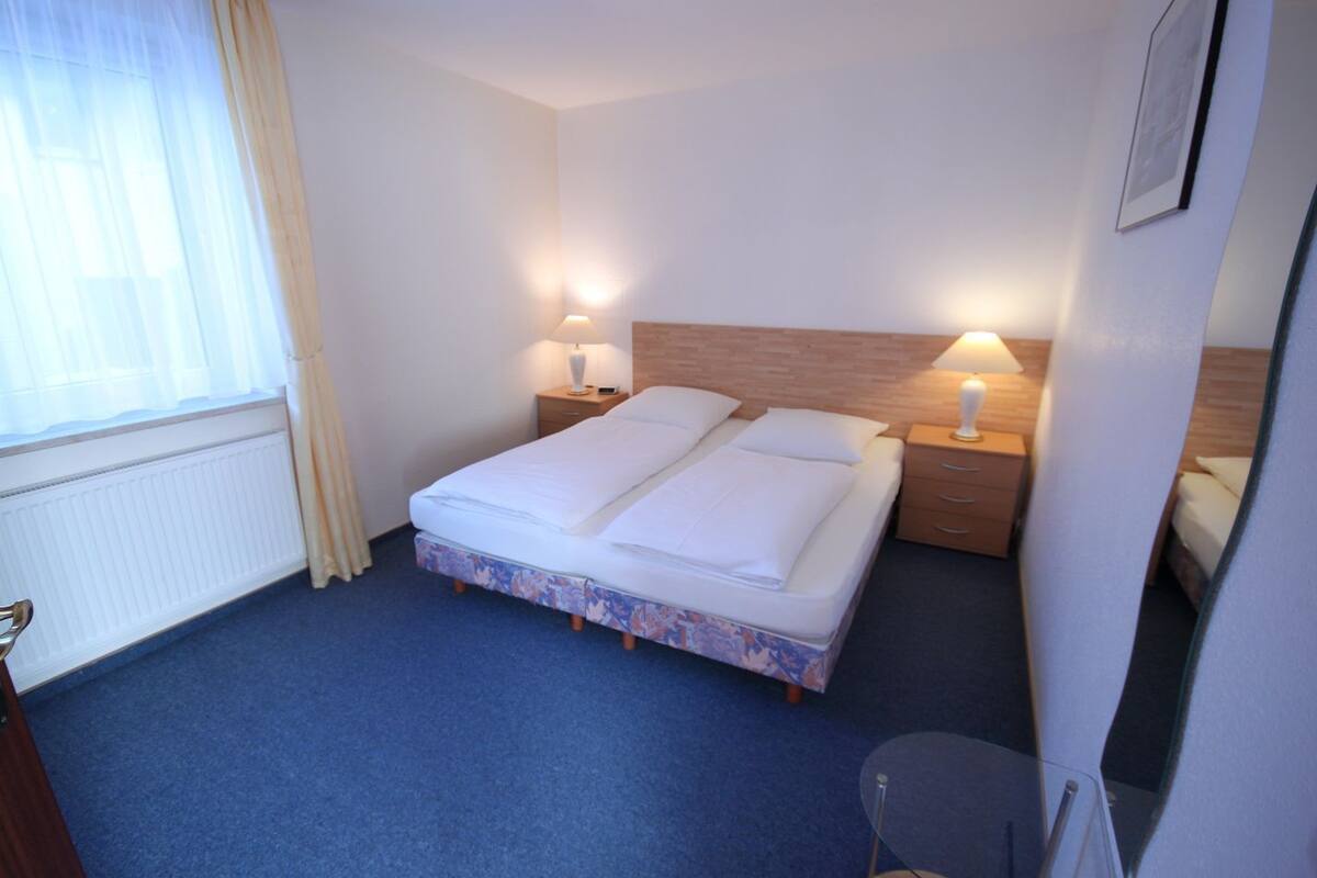 A hotel room with a bed, two bedside tables, a window with curtains, and a radiator.