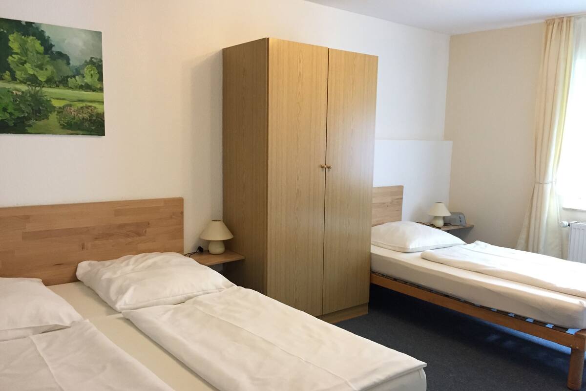 A hotel room with two single beds, a wooden wardrobe, a nightstand with a lamp, and a painting on the wall.