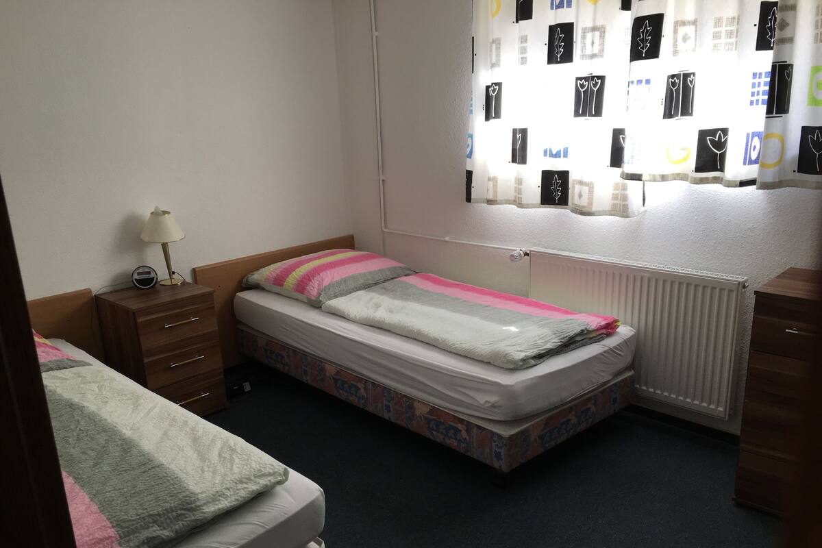 A bedroom with two single beds, a wooden dresser, a bedside table with a lamp, and a window with patterned curtains.