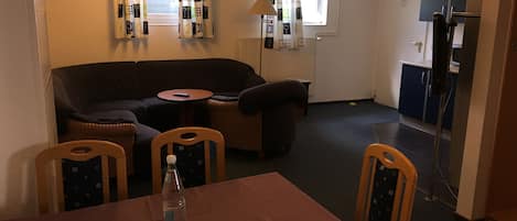 A cozy living area with a dark-colored sofa, a small round table, and a dining table with four chairs. A bottle of water is on the dining table. The room has a window with curtains, a lamp, and a door leading to another room.