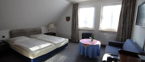 A hotel room with two beds, a small table with a tablecloth, a sofa, a TV, and a window with curtains.