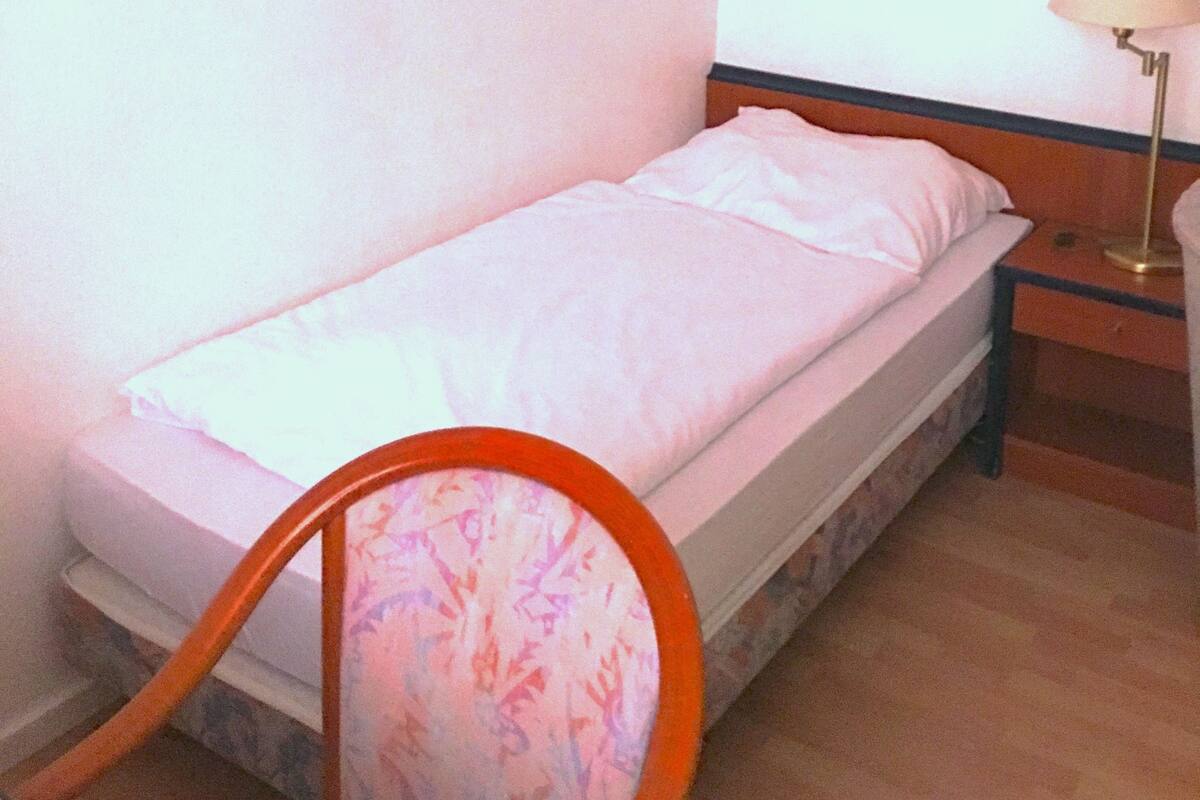 A single bed with a white mattress and pillows, a wooden nightstand with a lamp, and a wall with a landscape painting.