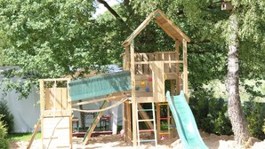 Una casita de juegos de madera con un tobogán y pared de escalar, rodeada de árboles y arena.